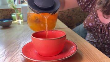 buckthorn tea from a glass teapot is poured into a bright pink mug on a plate wooden table Girl in a brown dress unrecognizable people tasty drink benefits vitamins video