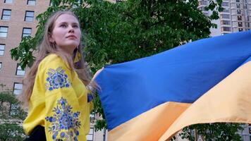 a beautiful Ukrainian woman in an embroidered holds Ukrainian flag on the background of the house a tree dreams of victory protection asks for help sad eyes fair hair confident strong imperious look video