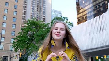 mignonne magnifique blond fille Jeune femme regards dans le Cadre et fait du une cœur avec sa mains joyeux dans une Jaune chemise sur une blanc vert Contexte rue l'amour paix dans Ukraine joie calme video