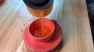 Meer Sanddorn Tee im ein dunkel Rosa Tasse. gießen von ein Glas Teekanne und trinken im ein gemütlich Cafe beim ein hölzern Tisch. video