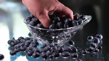 een vrouw picks groot vers bosbessen met haar hand, verstrooiing hen Aan de tafel in een glas bord, controle kwaliteit van de Product, voorbereidingen treffen de vulling voor taart, vitamines nuttig voor behandelen ogen video