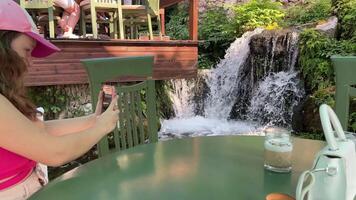 ung Tonårs flicka skjuter en på telefon för social nät av vattenfall i Albanien, två glasögon Coffee tabell, en keps, turist kläder, en promenad, en semester i de bergen i en Kafé video