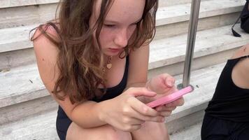 ung flicka Sammanträde på de steg och spelar på de telefon ser för Wi-Fi social nät telefon missbruk tonåringar ungdom svart minimal Kläder topp och shorts halsband med hjärtan europeisk video