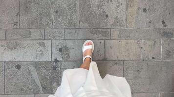 Beine von ein jung Mädchen Gehen entlang ein Stein Straße gefüttert mit Fliesen im Griechenland auf das Insel von Korfu ein Mädchen im ein Weiß Rock ein Frau langsam Spaziergänge entlang das Pflaster im Weiß Sandalen video