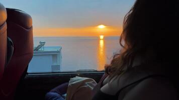 une fille avec duveteux cheveux regards dans le distance dans une autobus et regards à le le coucher du soleil magnifique vues Voyage par autobus adolescents ciel la nature video
