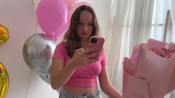 teenage girl celebrates birthday 15th anniversary balloons flowers numbers on white wall take picture for social networks on Internet holiday decorate room decorations jeans in pink style blouse video