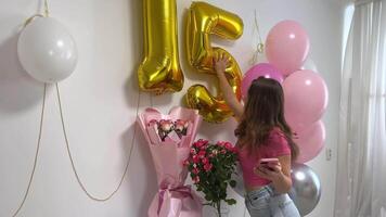 teenage girl celebrates birthday 15th anniversary balloons flowers numbers on white wall take picture for social networks on Internet holiday decorate room decorations jeans in pink style blouse video