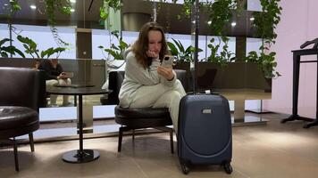 ragazza, valigia, telefono, aeroporto donna in attesa nel moderno aeroporto per sua ritardato volo con gambe su il grande rosso bagaglio utilizzando smartphone telefono in attesa per nuovo partenza volte video