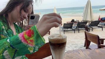 nehmen ein Foto von das Latté. Mädchen nimmt auf Handy, Mobiltelefon Telefon, Rühren ein Latté Eis Latté Cappuccino auf das Strand, ein jung Frau im ein Grün Pareo Blogs auf Sozial Netzwerke von das Meer, Ozean, Urlaub. video