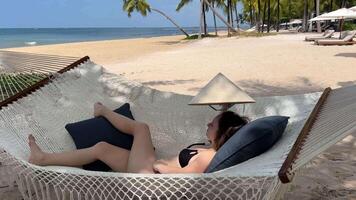 un niña mentiras en un hamaca con un vietnamita sombrero en su manos costoso hotel viaje anuncio mujer niña en un blanco pareo negro descansando mira a el teléfono usos móvil redes en el mar Oceano video