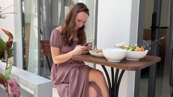 almoço para jovem mulher em terraço do hotel bangalô usando Móvel telefone em social redes dentro frente do a mesa lá estão branco pratos com Comida e pauzinhos oriental cozinha, Ásia período de férias turismo. video