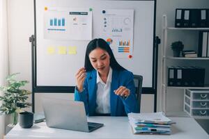 asiático mujer sensación migraña cabeza tensión.con exceso de trabajo mujer de negocios financiero mientras trabajando en ordenador portátil y tableta a moderno oficina. foto