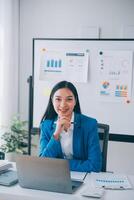 una alegre mujer de negocios que trabaja en una laptop en la oficina, una feliz y hermosa mujer de negocios asiática con traje formal trabaja en el lugar de trabajo. atractiva sonrisa de empleada de oficina. foto