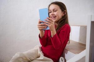 atención en un moderno azul teléfono inteligente en el manos de un borroso sonriente pequeño niño chica, teniendo divertido acecho , jugando virtual juegos en línea. digital adiccion. social medios de comunicación fanático niños foto