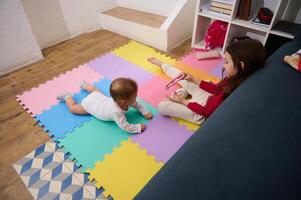 gastos generales ver de un pequeño niño chica, amoroso cuidando hermana jugando con su bebé chico hermano en un vistoso rompecabezas alfombra a acogedor hogar interior. niños. contento familia y mundo para niños día concepto foto