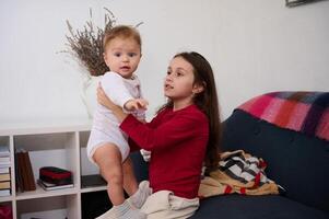 Cute sister holding her brother, an adorable baby boy on white bodysuit, playing together at home. Family relationships and love concept. Little child girl 6 years old helps her mom with a baby boy photo