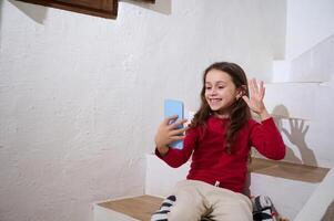 Cute little kid girl greeting friend or relatives having call on smartphone, smiling talking online sitting on steps at cozy home interior against white wall background. People and communication photo