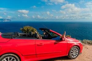 A woman is laying on her feet in a red car, looking out the window at the ocean photo