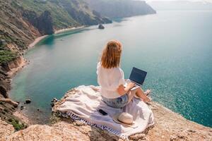 Lanza libre mujer trabajando en un ordenador portátil por el mar, mecanografía lejos en el teclado mientras disfrutando el hermosa vista, destacando el idea de remoto trabajar. foto