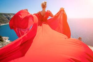 mujer mar rojo vestido. rubia con largo pelo en un soleado costa en un rojo fluido vestido, espalda vista, seda tela ondulación en el viento. en contra el fondo de el azul cielo y montañas en el costa. foto