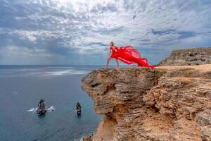 un mujer en un rojo seda vestir soportes por el océano, con montañas en el fondo, como su vestir se balancea en el brisa. foto