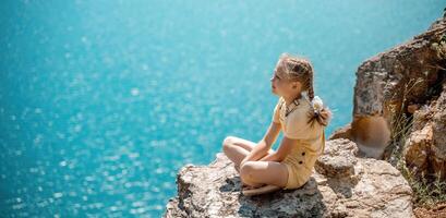 Happy girl perched atop a high rock above the sea, wearing a yellow jumpsuit and braided hair, signifying the concept of summer vacation at the beach. photo