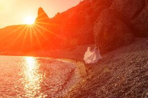 Mysterious woman silhouette long hair walks on the beach ocean water, sea nymph wind listens to the wave. Throws up a long white dress, a divine sunset. Artistic photo from the back without a face