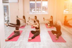 A group of six athletic women doing pilates or yoga on pink mats in front of a window in a beige loft studio interior. Teamwork, good mood and healthy lifestyle concept. photo