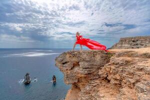 un mujer en un rojo seda vestir soportes por el océano, con montañas en el fondo, como su vestir se balancea en el brisa. foto