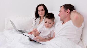 husband wife in bed with little son looking at tablet cartoon interesting game pictures laughing talking communication real people at home love life joyful people space for text ad social relations video