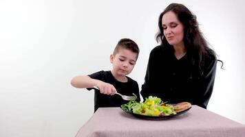 Mama und Sohn im schwarz Kleider beim Tabelle Junge Einspeisungen Mama mit Gemüse Salat Pflege Portion Eltern Vergnügen von kommunizieren mit Kinder Salat und Fleisch auf ein schwarz Teller Lachen Freude von Kommunikation video