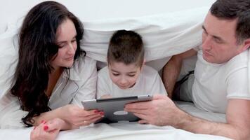 gente, familia y tecnología concepto madre, padre y pequeño chico con tableta ordenador personal computadora en cama a hogar o hotel habitación. contento familia gastar hora juntos acostado debajo blanco cobija en cama a dormitorio video