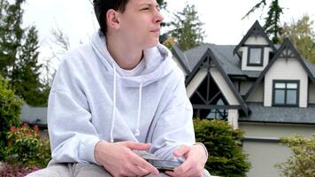 adolescent garçon dans une gris sweat-shirt séance avec une téléphone dans le sien mains sur le rue pique-nique attendre pour copains étreint une oreiller privé trimestre attendre à Regardez à le côté élever le sien tête ennuyé video