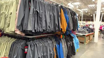 costco de gros achats Chariot une lot de le même vêtements dans différent tailles dans une supermarché une femme choisit une chemise à la recherche à il sur une cintre avec une Chariot dans sa mains et une chapeau cette elle acheté video