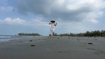 europeo niña vistiendo vietnamita sombrero joven mujer en un vacío playa en un blanco vestir camina a lo largo el arena participación su zapatos, Disparo en rojo épico video