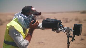 een cameraman in een reflecterende hesje is aan het kijken de het schieten in de woestijn. film schieten. de werk van een regisseur in moeilijk voorwaarden video
