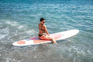 mujer savia mar. Deportes niña en un tabla de surf en el mar en un soleado verano día. en un negro baños traje, él se sienta en un sapa en el mar. descanso en el mar. foto