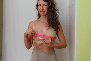 Cropped view of slim woman measuring breasts with tape measure at home, close up. Unrecognizable European woman checks the result of a weight loss diet or liposuction indoors. Healthy lifestyle. photo