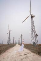 A woman in a white dress is walking down a dirt road in front of a row of wind turbines. photo