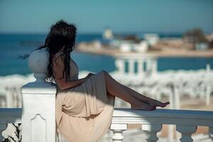 A woman in a tan dress is sitting on a white railing overlooking the ocean. The scene is serene and peaceful, with the woman enjoying the view and the calming sound of the waves. photo