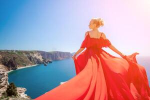 mujer mar rojo vestido. rubia con largo pelo en un soleado costa en un rojo fluido vestido, espalda vista, seda tela ondulación en el viento. en contra el fondo de el azul cielo y montañas en el costa. foto