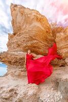 mujer rojo seda vestir soportes por el océano, con montañas en el fondo, como su vestir se balancea en el brisa. foto