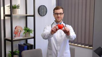 Doctor in white coat and glasses holding a red heart at hospital office. Medical health care and doctor staff service concept video