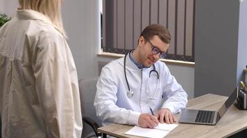 A young attractive doctor is having a consultation in his office. The doctor is making notes on the laptop, and the patient is standing by the table, smiling video