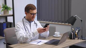 Male doctor medical worker in modern clinic wearing eyeglasses and white coat uniform using cell mobile smartphone apps, sitting at laptop computer. Medicine technologies health care concept video