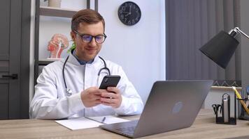 manlig läkare medicinsk arbetstagare i modern klinik bär glasögon och vit täcka enhetlig använder sig av cell mobil smartphone appar, Sammanträde på bärbar dator dator. medicin teknik hälsa vård begrepp video