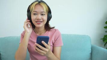 femme est séance sur une canapé et à la recherche à sa téléphone. elle est portant écouteurs et elle est écoute à la musique video