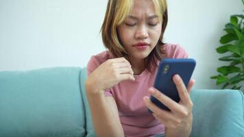 femme est séance sur une canapé et à la recherche à sa cellule téléphone. elle est portant une rose chemise et a sa main sur sa menton video