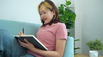 woman is writing in a notebook while sitting on a couch. She is wearing a pink shirt and has a pen in her hand. The scene suggests that she is taking notes or jotting down her thoughts video