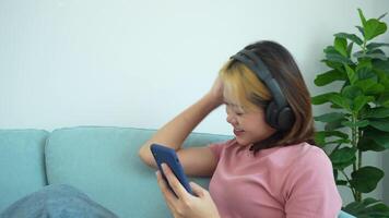 femme est séance sur une canapé et à la recherche à sa téléphone. elle est portant écouteurs et elle est écoute à la musique video